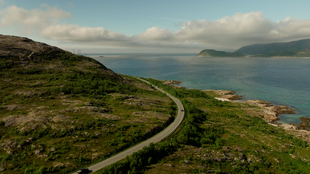 Aerial view of cars tracing a curving coastal road on Kvaloya island in Troms, with fjord water and rocky shoreline under soft clouds on a bright summer day