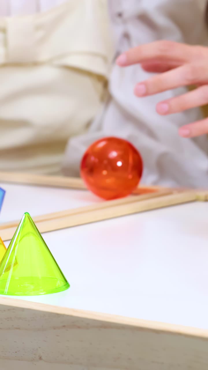 Lapset ja opettaja tutkivat värikkäitä läpinäkyviä geometrisia palikoita luokkahuoneen pöydällä lähikuvakameran liikkeellä.