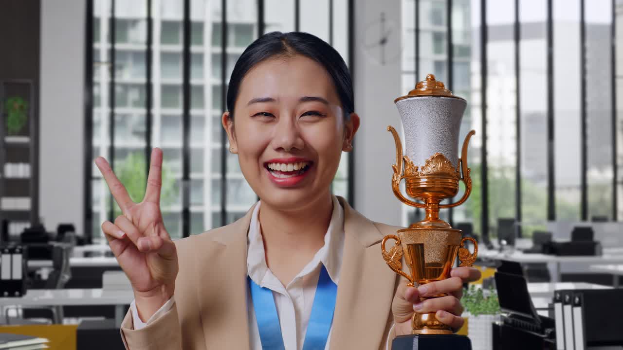 Close Up Of Asian Business Woman In A Suit With A Gold Medal And Trophy Showing Peace Gesture And Smiling To Camera As The First Winner In The Office