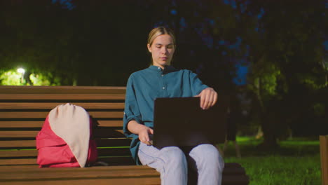 una mujer joven se sienta en un banco al aire libre por la noche, abriendo una computadora portátil bajo una iluminación suave, una bolsa roja descansa a su lado, rodeada de vegetación del parque, árboles y un poste de luz en el fondo