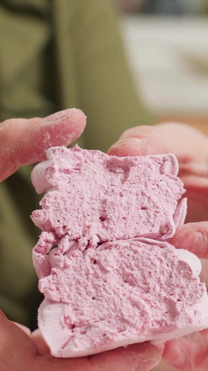 Primo piano di mani che manipolano materiale rosa friabile, rivelando. Mani coperte di polvere bianca fine lavorano per dividere l'oggetto. Sfondo interno sfocato con abiti verdi visibili.