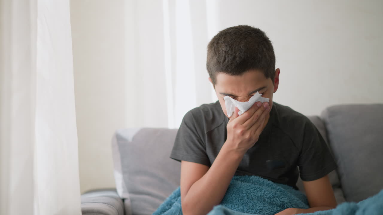 Sick boy sneezing into napkin covered with blanket on couch, wiping nose with tissue while looking tired, showing cold symptoms near window with soft light filtering in