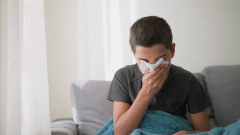 sick boy sneezing into napkin covered with blanket on couch, wiping nose with tissue while looking tired, showing cold symptoms near window with soft light filtering in