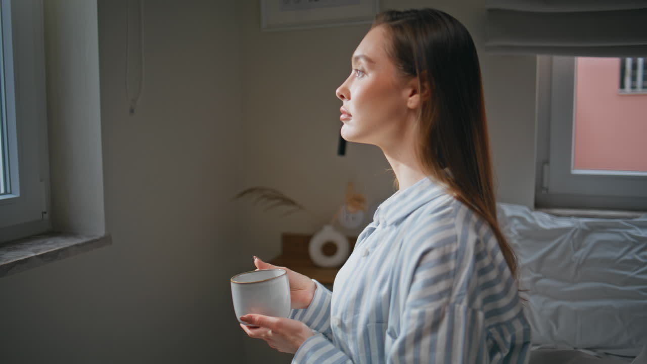 Serene woman looking window in bedroom holding coffee mug at tranquil morning