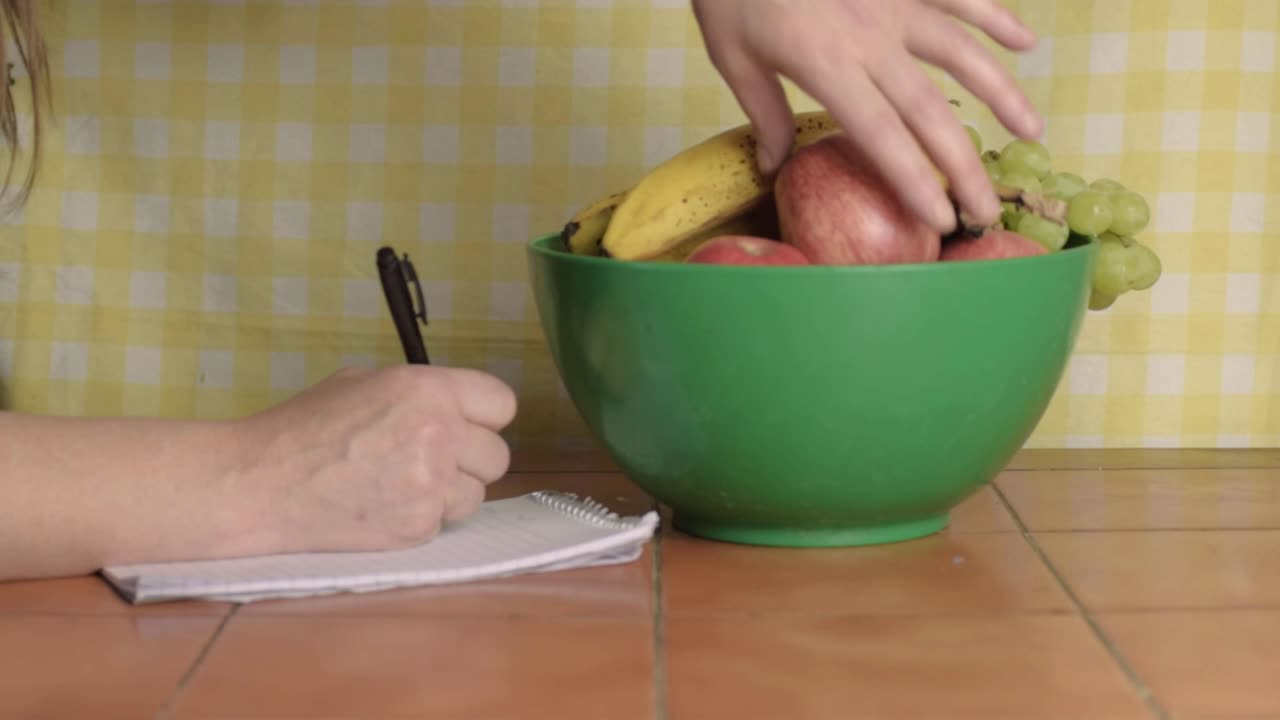 Woman writing at kitchen table with bowl of fruit