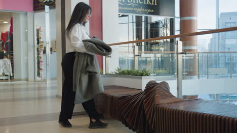 young girl holding jacket and book walking inside shopping mall approaches decorative bench surrounded by indoor plants, preparing to place her clothing item neatly in modern interior setting