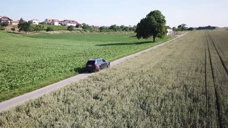 a volvo xc90 car drives alone on a road surrounded by agriculture fields in the swiss countryside, in the background some trees and a village, vaud, aerial drone shot