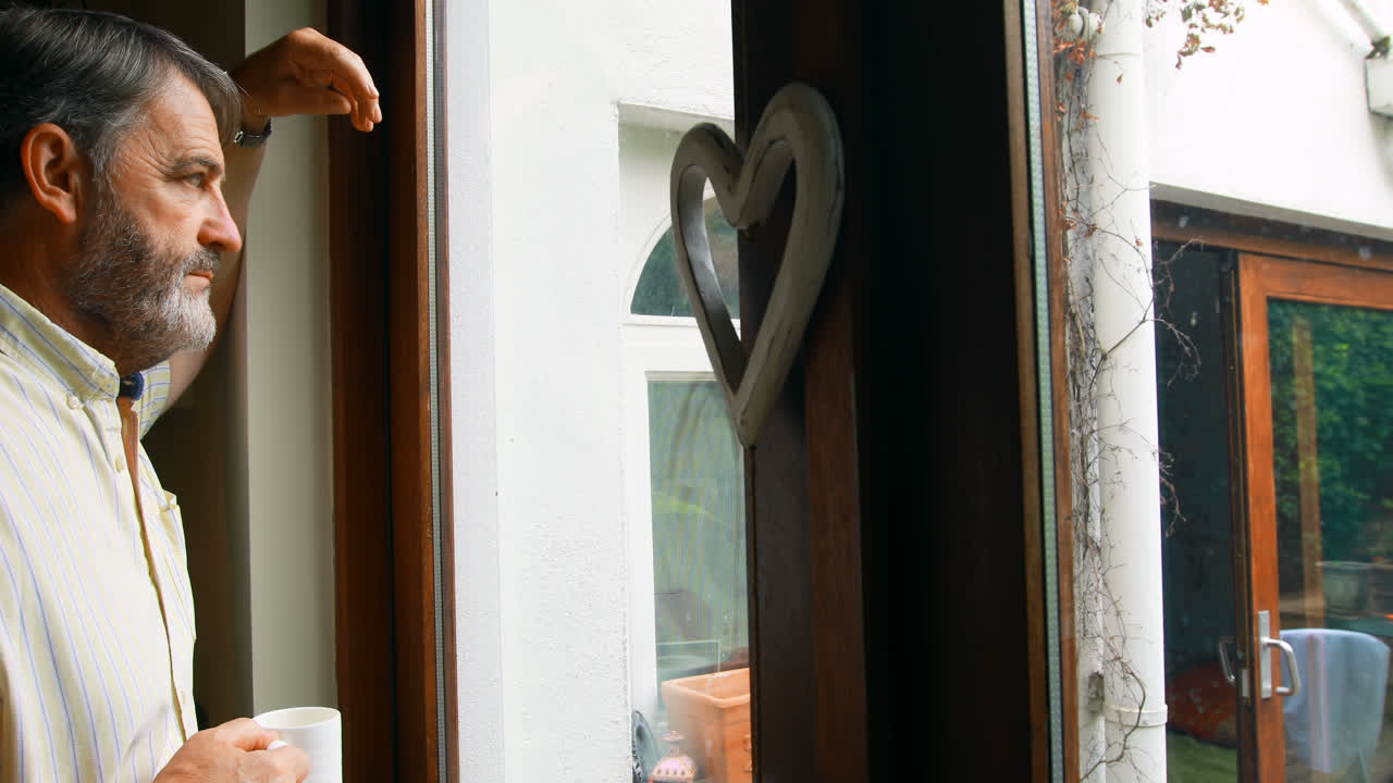 Senior man looking through window while having coffee at home 4k