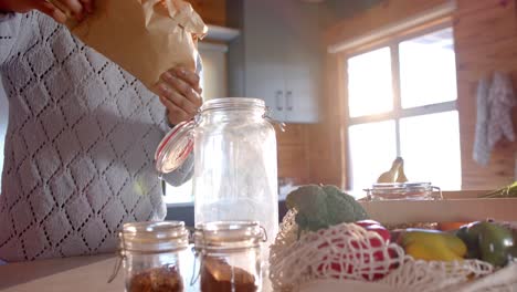 mid section of african american woman pouring pasta into jar in sunny kitchen, in slow motion