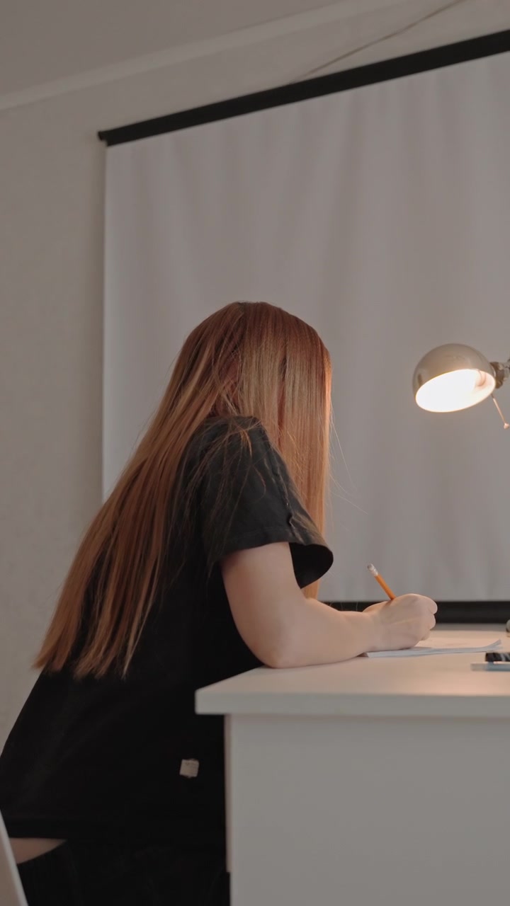 Young woman with long reddish brown hair writes. Desk holds papers and writing materials including orange pencil background is plain. Lamp provides focused warm illumination