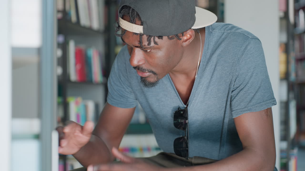 young student in cap squats in library aisle, leaning toward shelf while observing book titles, focused search for right volume under soft indoor light