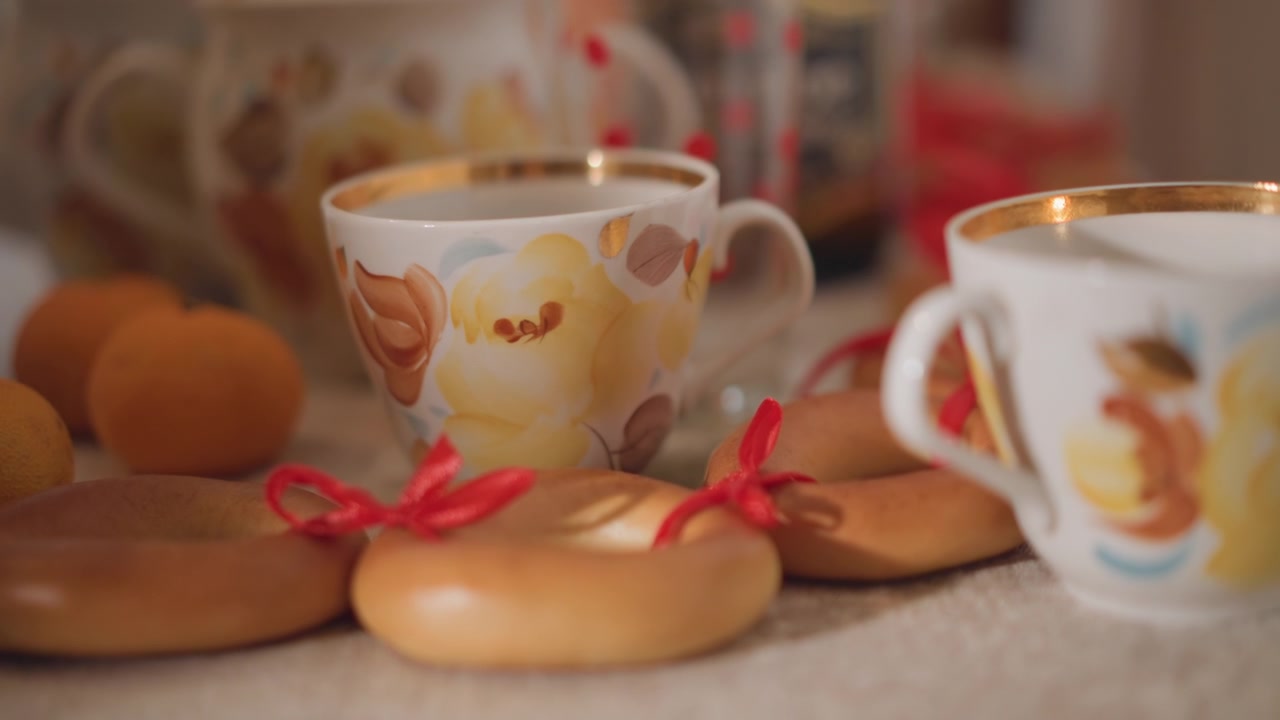 Closeup porcelain cups with ribboned biscuits, small mandarins and vintage china at cozy table, golden rim detail, soft warm light, holiday snack spread, intimate still life composition