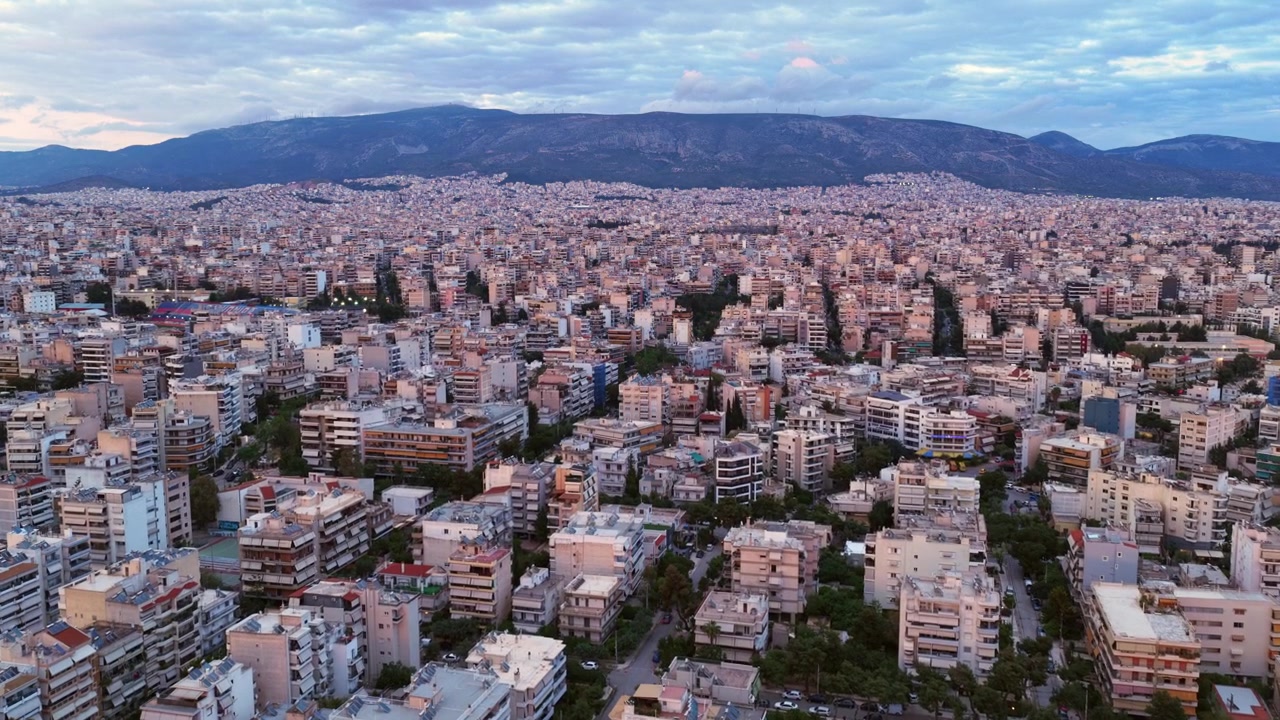 Ampia veduta aerea di Atene, Grecia, che mostra la densa espansione urbana di edifici residenziali con l'iconico Monte Imetto che si erge sullo sfondo sotto un cielo serale nuvoloso.