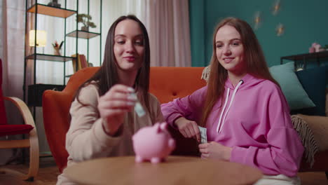 Girls-friends-siblings-sitting-on-floor-and-take-turns-dropping-dollar-banknote-into-piggy-bank