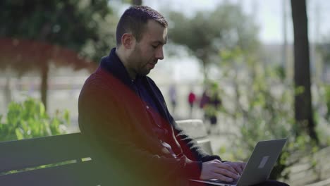 un hombre guapo y alegre escribiendo en una computadora portátil mientras está sentado en un banco