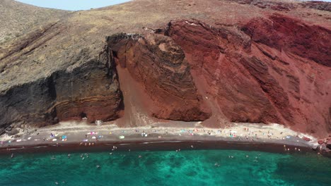 Aéreo-De-Arriba-Hacia-Abajo-Que-Revela-Una-Playa-Roja-Con-Agua-Turquesa,-Montañas-Y-Arena-De-Color-Rojo-En-Santorini,-Grecia