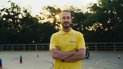 a male driving instructor in a yellow t-shirt with a beard and mustache folded his arms over his chest and poses