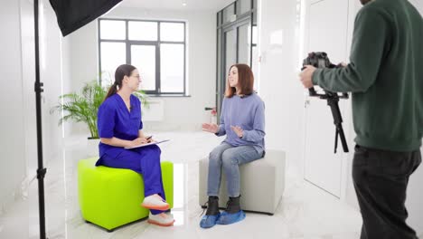 una chica morena segura con un suéter azul está entrevistando a una chica médica morena con gafas en un uniforme de otoño en una clínica moderna y brillante esto está siendo filmado por un tipo de prensa