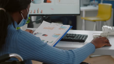 trabajadora negra paralizada con máscara protectora comprobando informes en la computadora