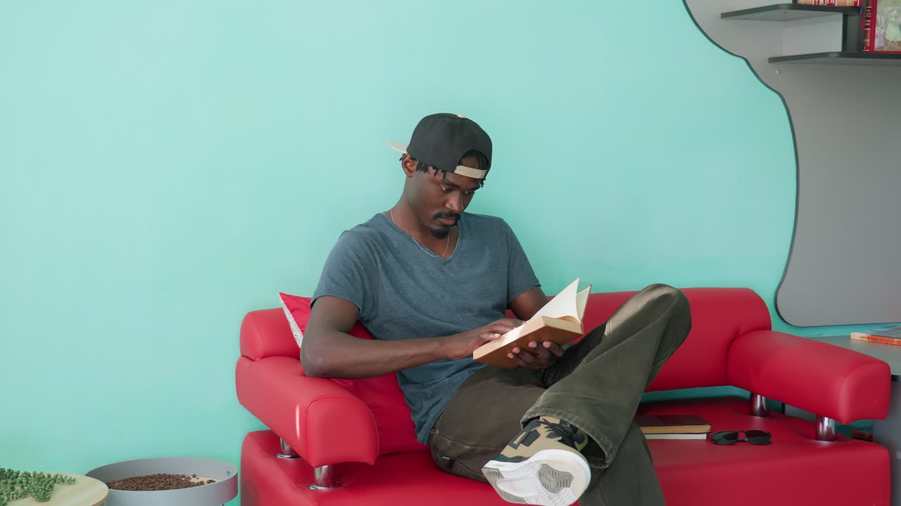 Jovem de boné sentado em um sofá vermelho em um espaço de leitura moderno e iluminado, abrindo um livro para ler enquanto coloca outro no sofá, com uma estante decorativa e livros coloridos ao fundo