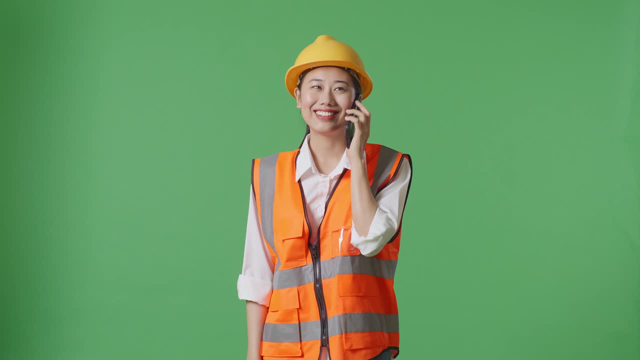 ingeniera asiática con casco de seguridad hablando en teléfono inteligente mientras está de pie en el estudio de fondo de la pantalla verde