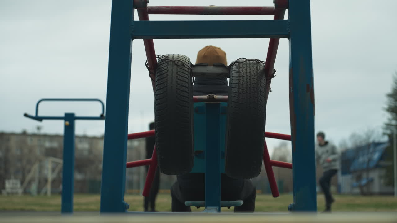 Back view of a coach stretching his hands out on workout equipment with two boys in the background, one in a gray sweater working out, with someone passing on an electric bike