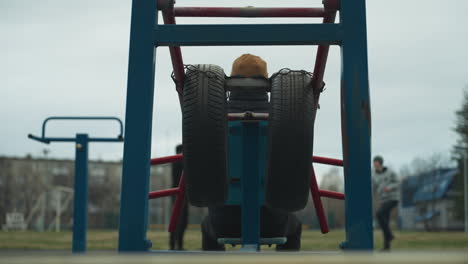 back view of a coach stretching his hands out on workout equipment with two boys in the background, one in a gray sweater working out, with someone passing on an electric bike