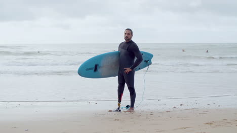 long shot of a handsome man in wetsuit with bionic leg standing on seashore with surfboard and smiling at the camera