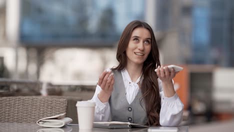 Portrait-of-a-smiling-business-woman-talking-on-the-phone-sitting-on-the-street-at-a-table