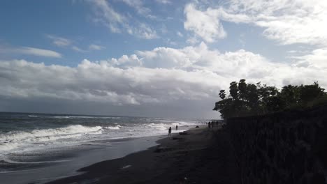 Acantilado-De-Arena-Negra-Y-Agua-De-Mar-En-Saba-Beach-Blahbatuh,-Gianyar-Bali-Indonesia,-Gente-Con-Los-Pies-En-La-Orilla-Del-Océano,-Hora-De-La-Tarde,-Cielo-Azul