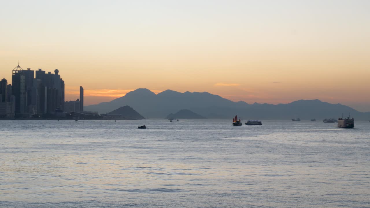 Sunset over a city harbor with a skyline, mountains, and boats on the water
