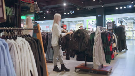 expectant mother with long silky hair and black handbag lifts winter coat from clothing rack inside boutique as shoppers pass by outside transparent store window in brightly lit modern shopping mall
