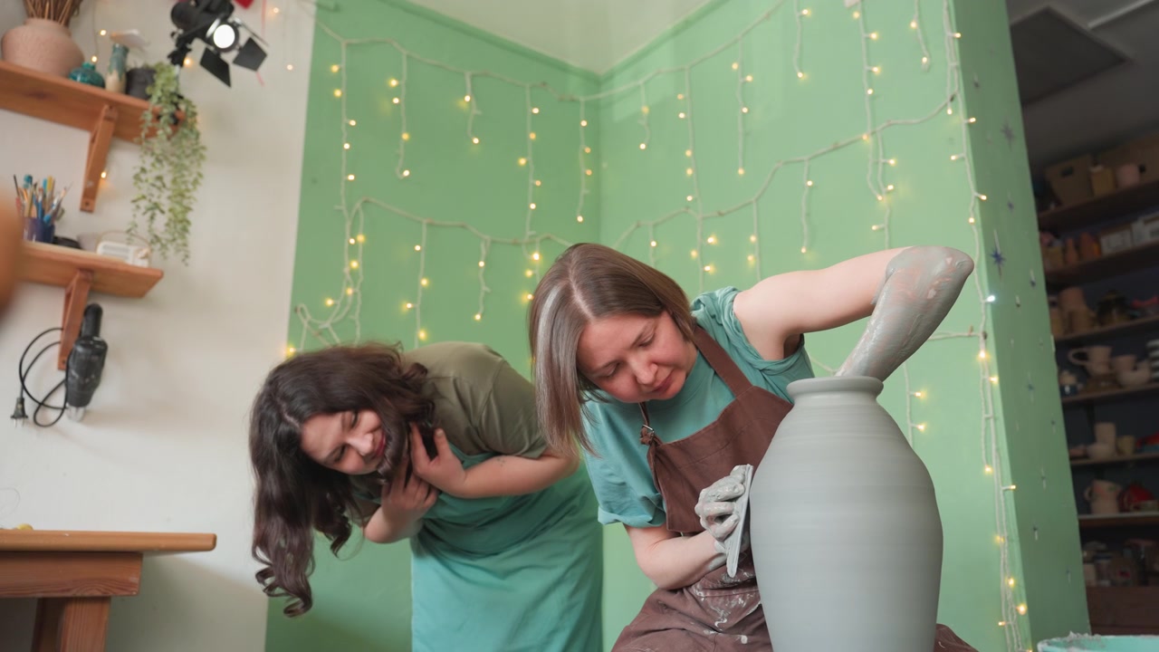 Two women in pottery workshop shaping clay vase, mentor guiding student at wheel, warm green studio backdrop with string lights and shelves, hands covered in wet clay, collaborative lesson
