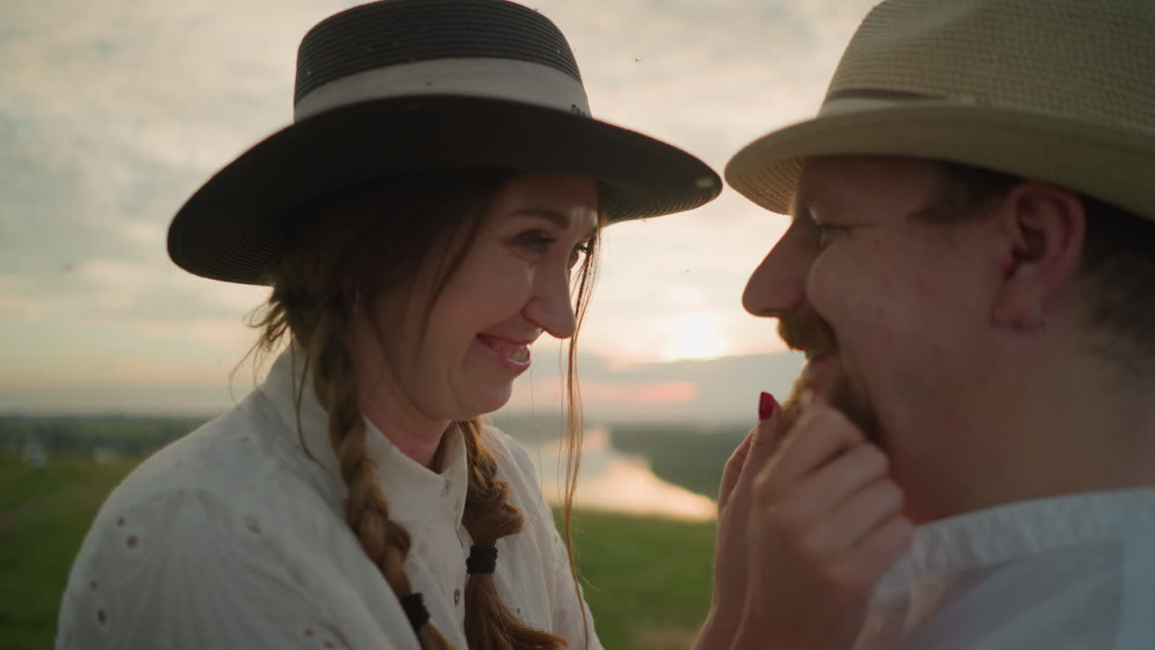 A wife in a white dress, a hat and bracelets, smiles lovingly as she touches her husband's face and beard in a grassy field at sunset. He wears a white shirt and a hat, and they share a joyful moment