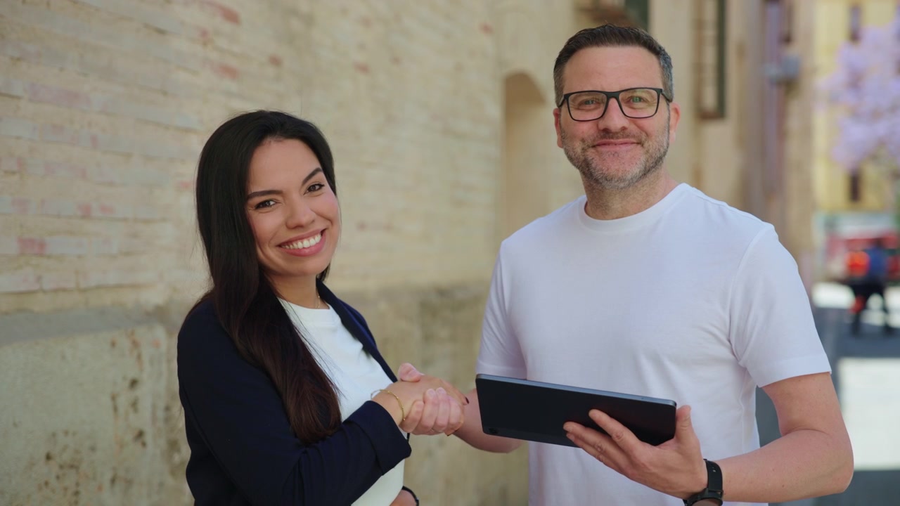 Un uomo d'affari e una donna d'affari esaminano insieme un tablet digitale all'aperto, poi si stringono la mano e sorridono direttamente alla telecamera per celebrare un affare di successo, registrato al rallentatore.