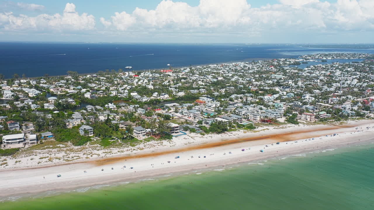 Anna Maria Island’s wide sandy shoreline meets turquoise waters, while residential homes and lush greenery stretch inland beneath a bright summer sky
