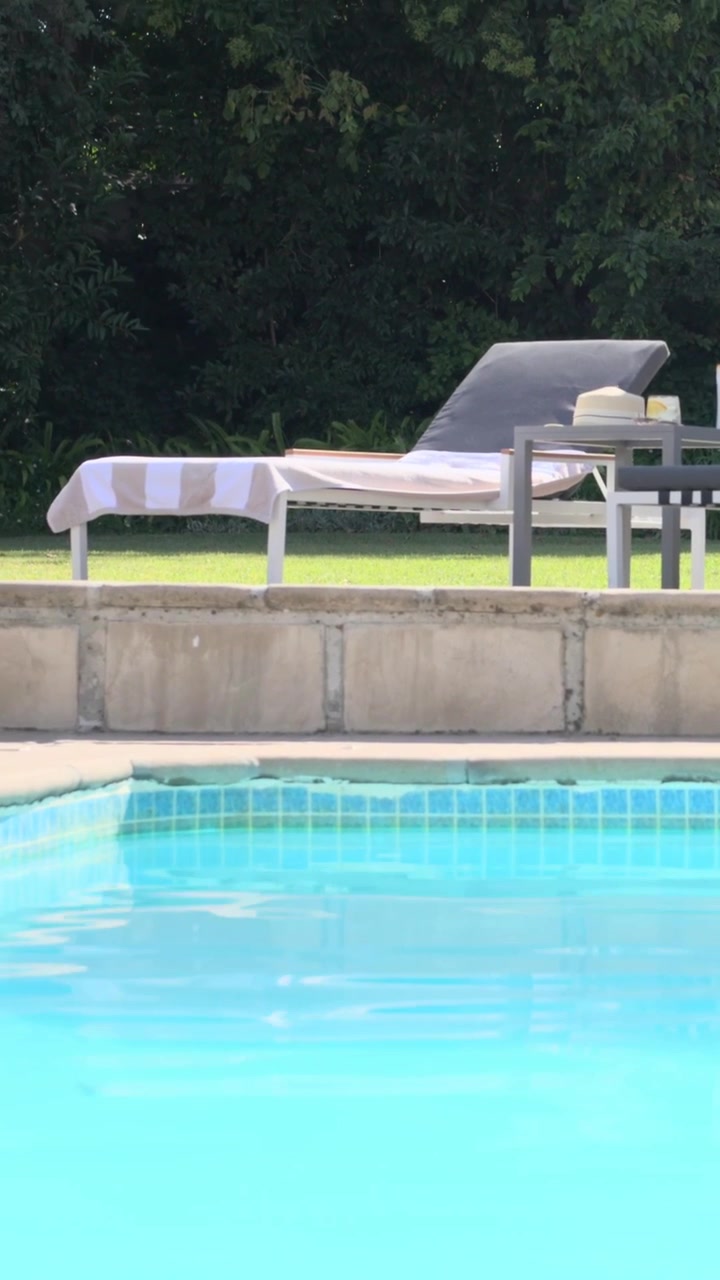 Vertikal video: Kamera panorerer fra blå pool, stiger op til dækket, afslører liggestole, håndklæde og hat