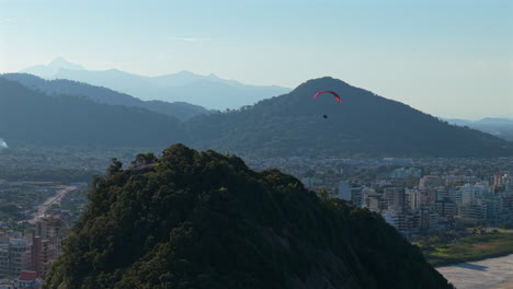 Drone panning shot tracking a paraglider above Caiobá, with a steep forested hill in the foreground, city buildings and smoky vegetation in the background