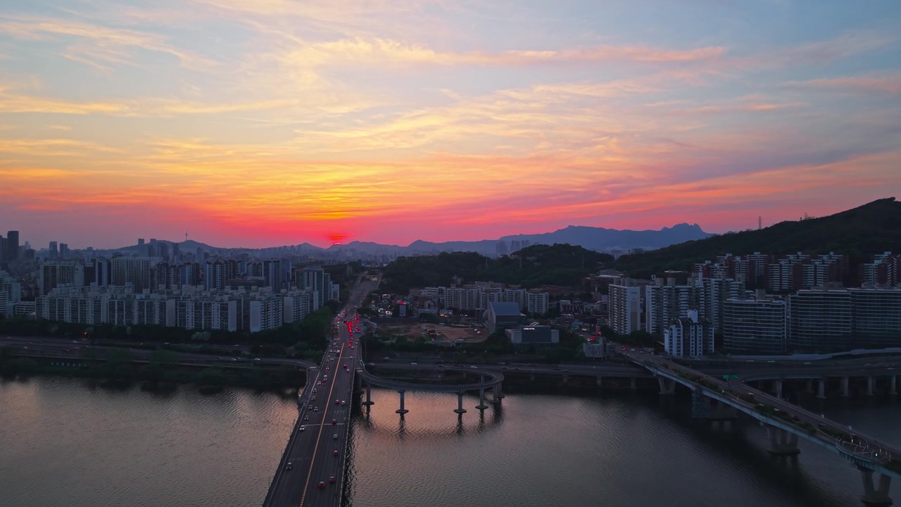 Volo aereo in avanti sopra il ponte Gwangjin con traffico serale verso un drammatico cielo rosso fuoco al tramonto sopra il fiume Han con il ponte Cheonho a Seul