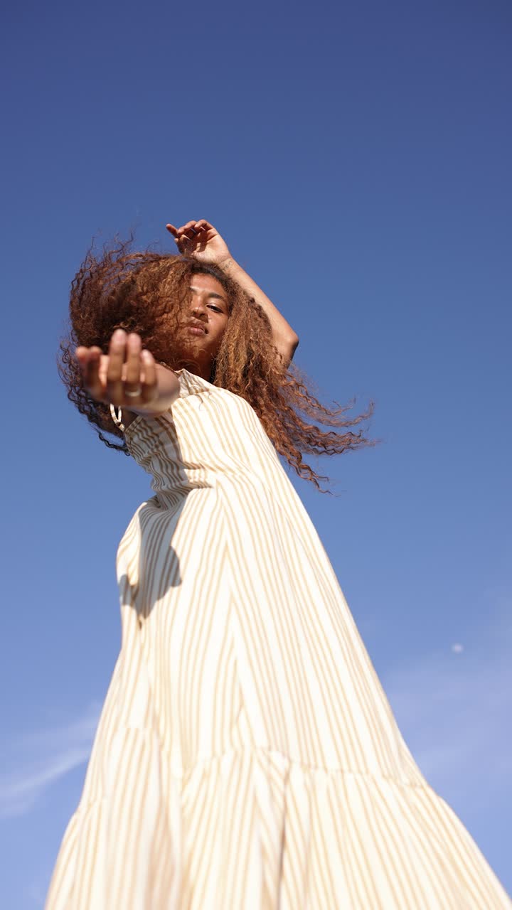 A young woman with curly hair poses against a clear blue sky, embracing the sun and wind