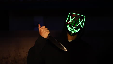 a terrifying killer with a knife and a generic scary halloween mask standing in the shadows before attacking his victim