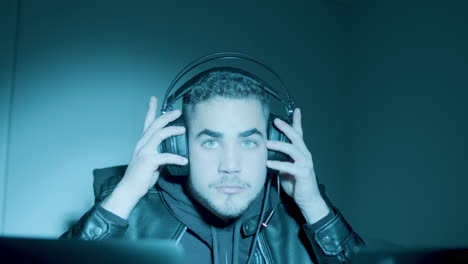 joven hacker caucásico sentado por la noche frente a la pantalla de un portátil, quitándose los auriculares y mirando la cámara