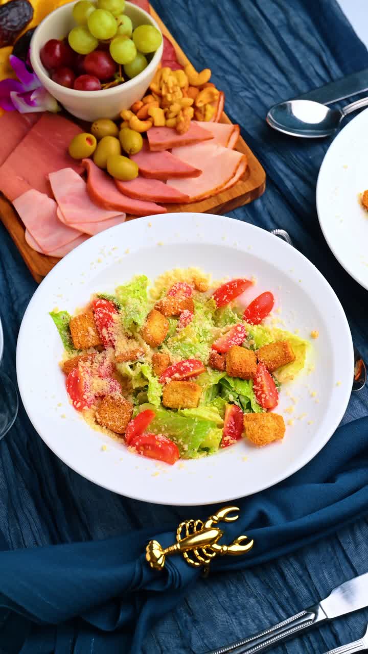 Insalata Caesar con crostini e pomodori accanto a un tagliere di salumi. Vista dall'alto di insalata Caesar e piatto di salumi su sfondo di tessuto blu testurizzato
