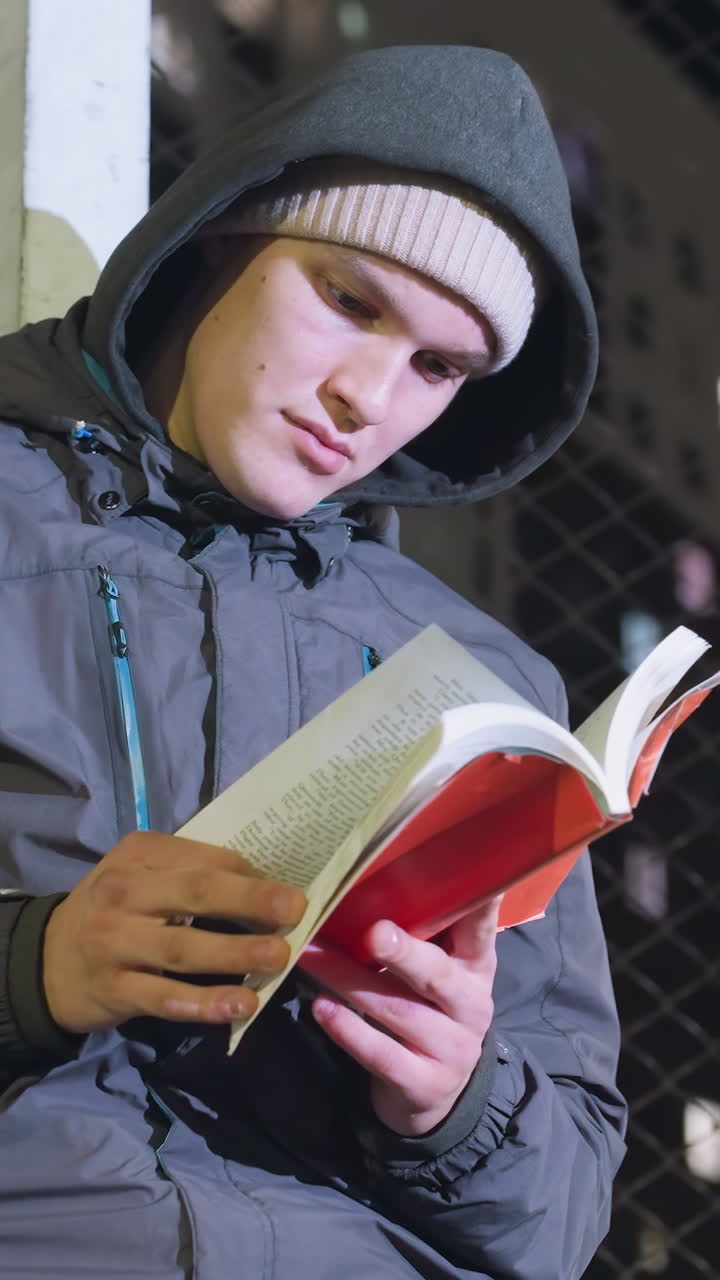 hombre con capucha y gorra apoyado contra un poste de meta metálico leyendo libro rojo al aire libre por la noche con valla de malla y fondo de ciudad urbana iluminado, volviendo páginas pensativamente