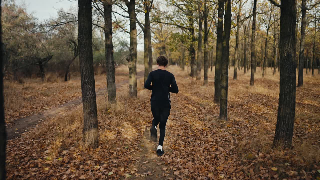 vista trasera de un feliz y confiado atleta masculino corriendo en un uniforme deportivo negro a lo largo de un camino de tierra en un bosque de otoño con hojas marrones caídas