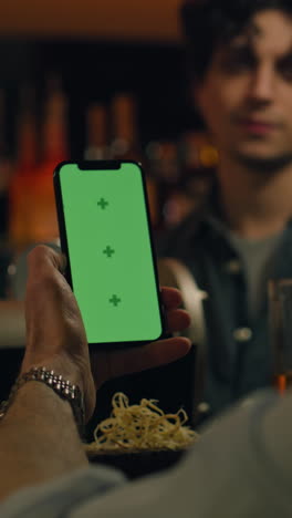 Vertical-shot-of-Mature-sports-fan-uses-mobile-phone-with-green-screen,-bets-on-football-match-online,-talks-with-bartender-and-friend.-Man-checks-bookmaker-ratings-in-app-sitting-at-bar-counter-in-pub.