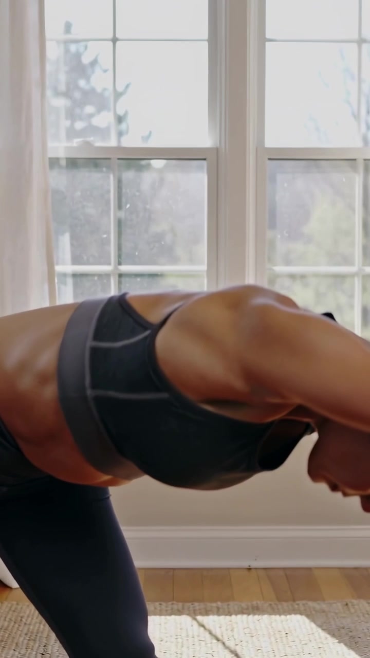 Video verticale: Iniziando con le braccia alzate, donna in reggiseno e collant che si piega per fare stretching vicino alla finestra