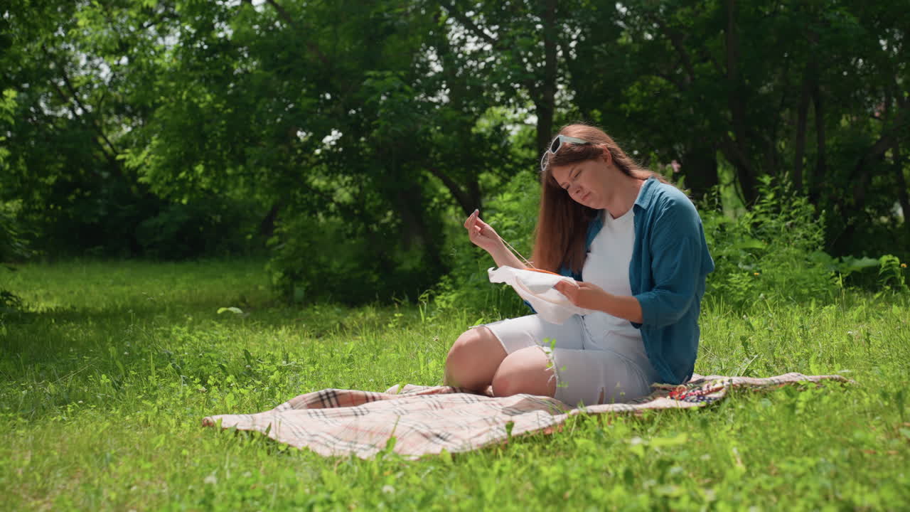 Young lady embroidering under sunlight while sitting on blanket in lush summer park surrounded by green trees, focused on delicate needlework, enjoying peaceful outdoor atmosphere