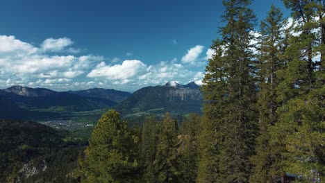 Wide-dreamy-and-epic-mountain-panorama-tree-fly-through-at-scenic-Bavaria-Elmau-castle-and-snowy-glacier-peaks-in-the-Bavarian-Austrian-alps-on-a-cloudy-and-sunny-day-along-forest-and-hills-in-nature