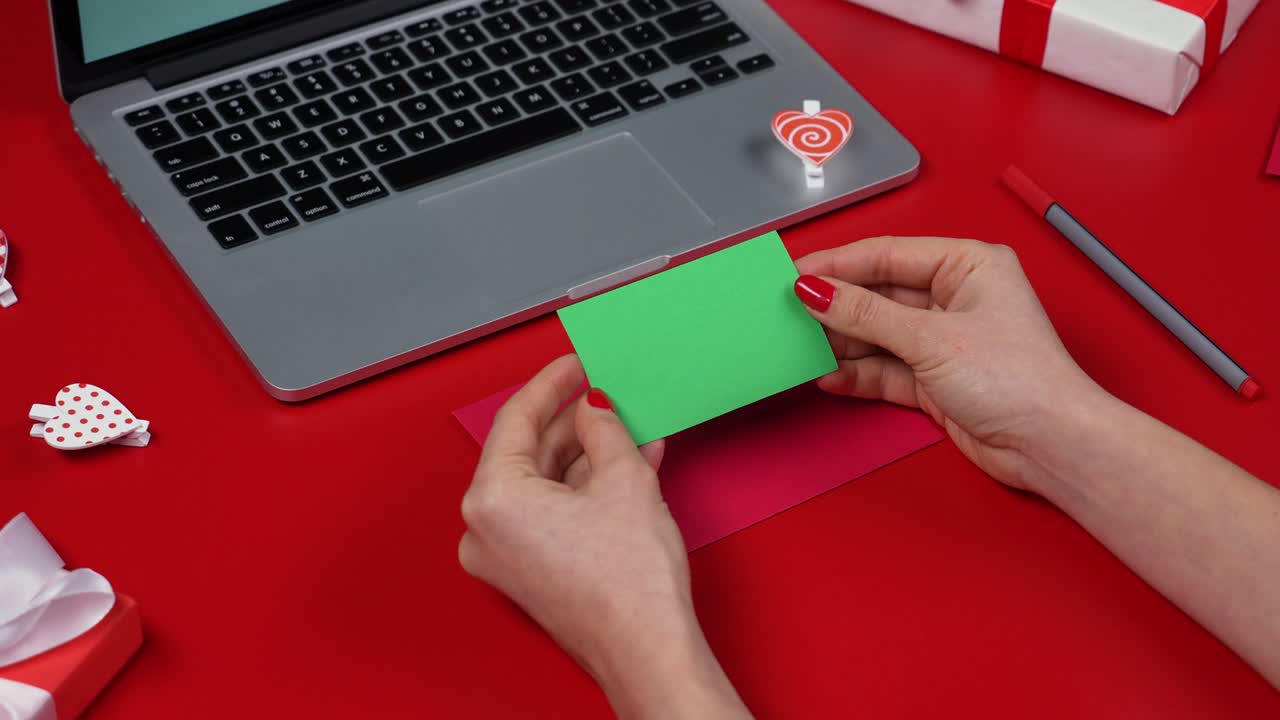 las manos de la mujer ponen una tarjeta postal de amor de san valentín con pantalla verde llave de croma en el sobre festivo. vista superior de la mesa roja con una computadora portátil y cajas de regalos. concepto del día de san valentine. cerca. cámara lenta